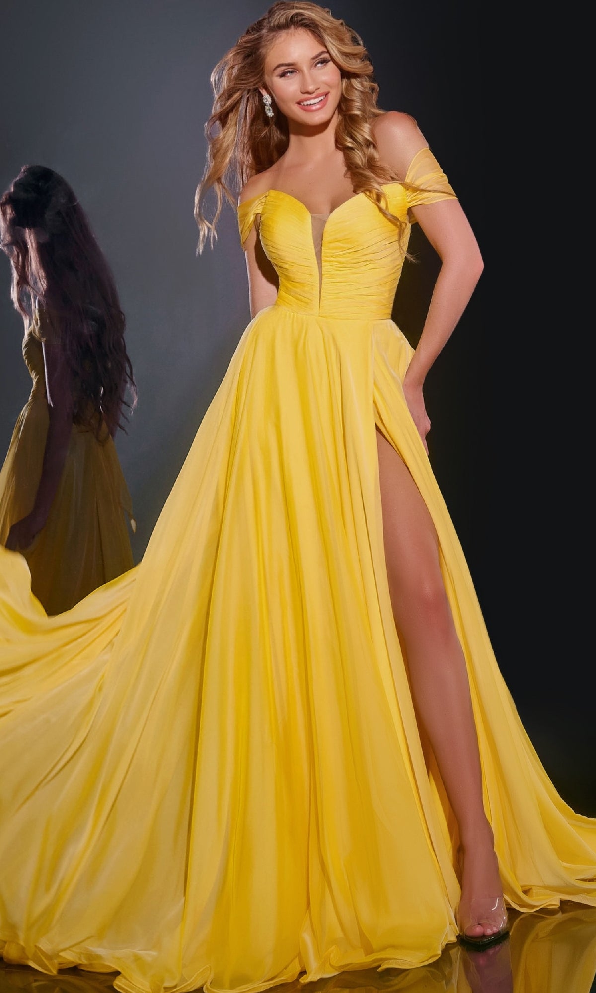 A woman in a yellow Long Chiffon A-Line Prom Dress (Jovani 43842) with an off-the-shoulder neckline and thigh-high slit poses smiling in front of a mirror, her wavy hair cascading over her shoulders.
